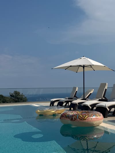 This media file depicts a sunny day at a poolside in Krimovice, Montenegro. The scene is dominated by a clear blue sky with a few wispy clouds, and a small bird is visible soaring in the distance. In the foreground, a bright white umbrella is open, casting a shadow on the surrounding environment. Next to the umbrella, a line of white lounge chairs with dark cushions are arranged, suggesting a relaxation area.
The main focal point in the water are two inflatable pool toys. One is a donut-shaped float adorned with a chocolate frosting design and colorful sprinkles. Adjacent to it is a clear inflatable ring, possibly for a drink or a smaller float. The swimming pool itself features a tiled bottom in a light blue hue. The water reflects the sky and the poolside furniture, creating a serene and inviting atmosphere. A glass barrier runs along the edge of the pool area, behind which trees and greenery are visible, hinting at a lush environment. The overall impression is one of a leisurely summer day, perfect for enjoying the water and sunshine. The time of day appears to be midday, given the bright sunlight and clear sky.