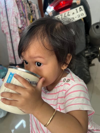 A young child is drinking from a metal mug. The child has dark hair and is wearing a pink and white striped shirt. A license plate with the numbers "CZL44 IA 3-28" is visible in the background, along with a motorcycle and clothing on a rack. The scene appears to be indoors, possibly a home, in Kecamatan Pati, Indonesia.