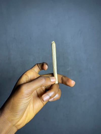 A close-up, waist-up, medium shot shows a person's dark-skinned hand holding a rolled cigarette between their thumb and index finger. The cigarette is pale yellow and slightly unrolled at the tip. The background is a dark, out-of-focus gray.