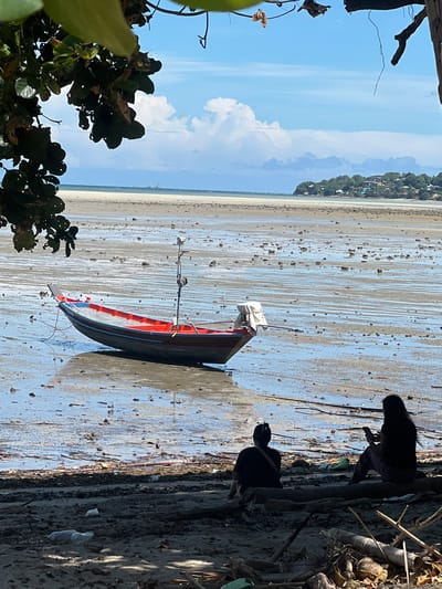 The image depicts a low tide scene on the coast of Koh Phangan, Thailand. A red and blue fishing boat is grounded on the wet sand, its hull reflecting the sky. The vast expanse of the receding tide reveals a muddy seabed littered with debris and small rocks. In the foreground, two individuals are seated on a log, facing the water. One person, in silhouette, is looking down at a phone, while the other person, also in silhouette, is sitting with their back to the viewer. Behind them, the shoreline is covered in sand, driftwood, and various forms of litter, including plastic bottles. In the distance, a treed hillside with buildings stretches along the coast, and the ocean meets a hazy horizon under a partly cloudy sky.