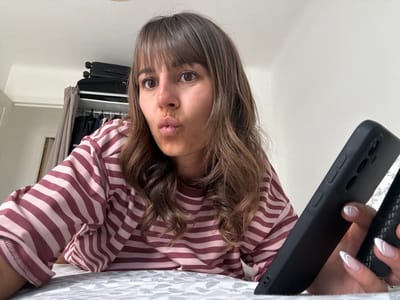 In an indoor setting, a young woman with brown hair and bangs is captured in a close-up, waist-up shot, looking directly at the camera with her lips pursed. She is wearing a horizontally striped long-sleeved shirt in shades of pink and maroon. Her right hand is holding a black smartphone, positioned in the foreground. The background reveals a closet with clothing and luggage, suggesting a bedroom or hotel room. The scene does not provide any specific geographical markers that would indicate the location is Cannes, France.