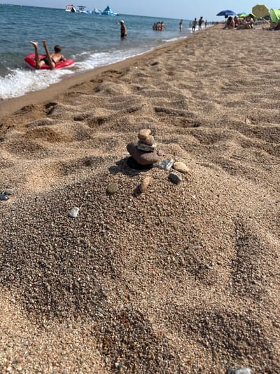 The image depicts a close-up view of a sandy beach in Blanes, Spain, with the sea and other beachgoers in the background. In the foreground, a small cairn of stones is balanced on a mound of sand. The sea is visible with a person in a red inflatable ring being splashed by waves. Further out, several inflatable water structures are present. Along the shoreline, people are visible, some sitting under umbrellas, while others are in the water. The sand appears coarse-grained and a mixture of light brown and tan hues. The sky is clear and blue, suggesting a sunny day.