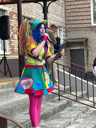 A clown in colorful costume stands on a metal platform in Dmitrov, Russia, holding a microphone and black clipboard. The clown has blue hair, pink tights, and a patchwork-style outfit with a "WOW!" patch. A man in a tan jacket and black bag lies on the ground behind the clown. A metal fire escape is behind them. The background consists of brick buildings.