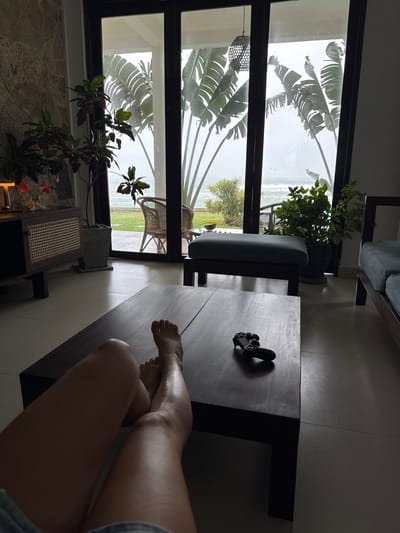 The image depicts a person's legs and feet resting on a dark wooden coffee table in an indoor setting, possibly a living room or lounge. The perspective is from the person's viewpoint, looking down and slightly to the side. The person is wearing denim shorts.
Beyond the coffee table, a large window reveals an outdoor scene. Lush, large-leafed tropical plants, likely banana trees or similar, dominate the view through the window, with their fronds casting shadows. Outside, the weather appears overcast and possibly rainy, with a pale, diffused light. Hints of a body of water and some greenery can be seen in the distance, suggesting a location near the coast or a tropical landscape. A wicker chair is visible outside, positioned on a balcony or patio.
Inside, near the window, are several potted plants, one with dark-red and green leaves, and another with vibrant green foliage. A decorative woven lamp shade hangs from the ceiling. On the coffee table, next to the person's legs, is a black video game controller. The overall atmosphere suggests a moment of relaxation and indoor leisure, with the overcast weather perhaps encouraging time spent indoors. The lighting is dim, with shadows cast by the furniture and plants. The location cues of tropical plants and the likely coastal view point towards Goviyapana, Sri Lanka. No visible text is present.