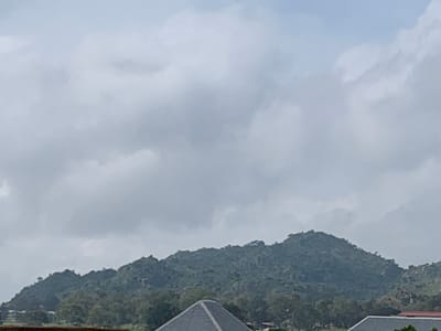 The image shows a cloudy sky above a verdant, hilly landscape. The hills are covered in dense vegetation, with some rocky outcrops visible. In the foreground, a dark, pyramidal roof structure is partially visible, suggesting the presence of buildings or structures. The overall scene evokes a natural, possibly rural or semi-urban environment. The city of Kuru Karama, Nigeria, is located in the Jos Plateau region, known for its hilly terrain and natural beauty, which aligns with the visual elements present in the image. The weather appears overcast, contributing to a muted color palette. There are no people or distinct actions depicted in the image.