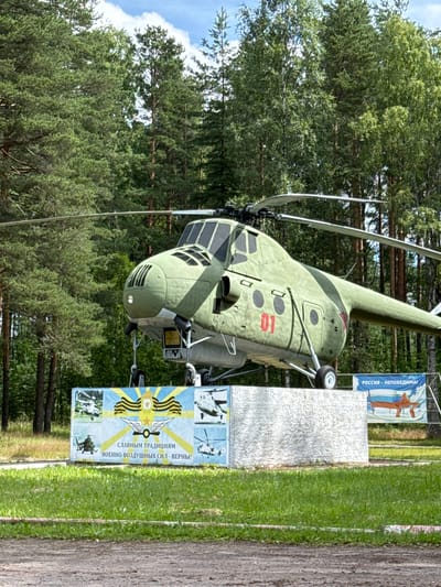 This is a medium shot, eye-level, outdoor, daytime photograph taken in the city of Pribylovo, Russia. The main subject is a green, vintage helicopter mounted on a concrete pedestal. The helicopter is olive green with a red number "01" on its side. It has a visible propeller on top and landing gear. The pedestal is covered with a white, textured material, and in front of it are two banners. The left banner features images of other aircraft and text in Russian, including "СЛАВНЫМ ТРАДИЦИЯМ" and "ВОЕННО-ВОЗДУШНЫХ СИЛ - ВЕРНЫ!". The right banner shows a stylized aircraft and the Russian text "РОССИЯ - НЕПОБЕДИМАЯ". The background consists of a dense forest of tall green pine and birch trees under a bright blue sky with scattered clouds. The ground in the foreground is a mix of dirt and green grass. There are no people visible in the frame.