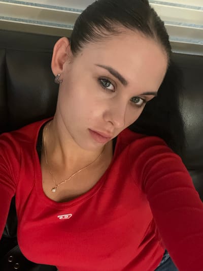 A female individual is depicted in a close-up, chest-up portrait. She has dark hair pulled back, light-colored eyes, and is wearing small, silver-colored hoop earrings. A thin, gold-colored chain necklace with a small, light-colored, possibly star-shaped or floral-shaped pendant rests on her sternum. She wears a long-sleeved, ribbed red top with a scoop neckline, featuring a small white "Diesel" logo on the upper left chest. A dark garment is visible beneath the red top's neckline. Part of a dark, textured surface, possibly leather upholstery, occupies the background behind her, suggesting an indoor setting. A dark object with silver-colored eyelets or rivets is partially visible in the lower left, and dark blue fabric, likely denim, is visible in the lower right. The specific location within Mapo-gu, South Korea, is undetermined based on visual information.