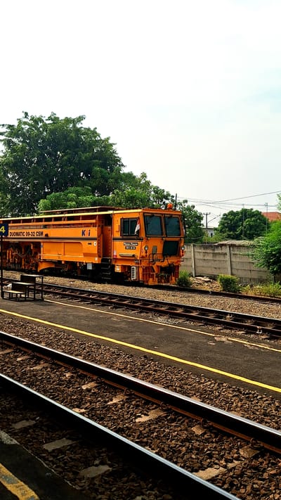 The image shows an orange rail maintenance vehicle, a Plasser & Theurer Duomatic 09-32 CSM, parked on a railway track in Karawang, Indonesia. The vehicle is positioned parallel to the tracks and adjacent to a platform. Beyond the tracks and vehicle, there's greenery, a concrete wall, and a section of sky. The platform edge is visible in the foreground.