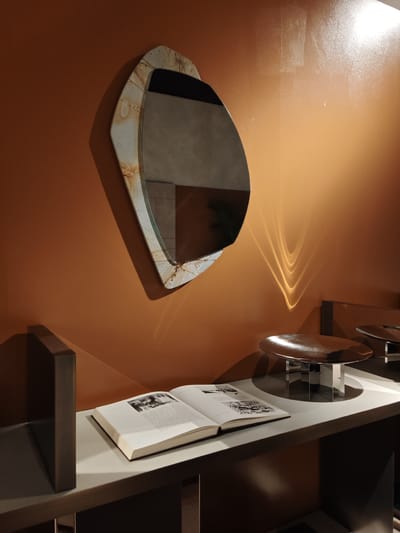 The image presents an interior scene, possibly a display at an event or exhibition in Rho, Italy. It features an orange-toned wall as a backdrop. A geometrically shaped mirror with a distressed frame is mounted on the wall. Below, a light-colored shelf supports a book opened to pages displaying images of what appears to be artwork or architecture. Nearby sits a sleek, shallow metallic bowl on a geometric pedestal, and another similar bowl can be partially seen in the background. The scene is illuminated, creating reflections and highlights, especially on the wall.