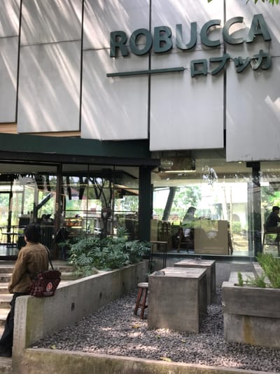 The image shows the exterior of a cafe called "ROBUCCA" in Malang, Indonesia, located at coordinates -7.98 latitude and 112.617 longitude. The cafe has a white facade with a large sign in green lettering. There are large glass windows, and the cafe has a patio area with concrete tables and chairs, as well as a small stone garden. A woman wearing a green jacket is standing near the entrance. The patio is partly shaded by the trees. The interior of the cafe is visible through the windows, showing several tables and chairs and some people sitting at them. There are green plants surrounding the cafe, adding to the ambiance. The image was taken on a sunny day.