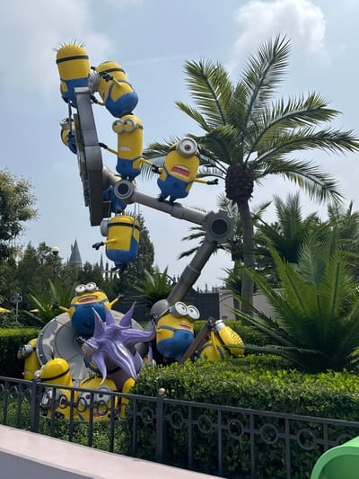 This is a medium shot of a themed amusement park attraction featuring Minion characters from the Despicable Me franchise. The attraction consists of several Minion figures, some appearing to be in motion, mounted on a central metallic structure. In the foreground, a black metal fence separates the viewer from the attraction, behind which is lush green shrubbery. In the background, palm trees and other foliage are visible, along with a hazy sky. A distinctive castle-like structure is visible in the far background, suggesting a larger themed environment. The specific location is identified as Tongzhou District, China.