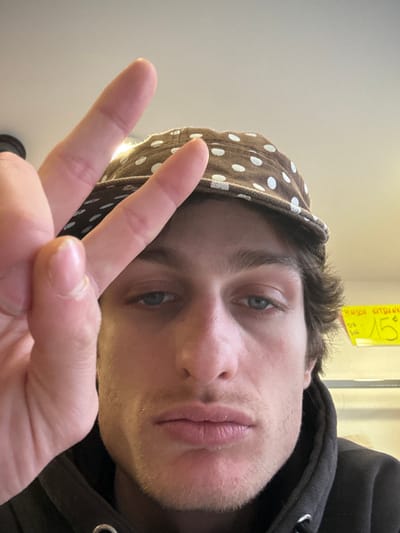 The image depicts a male individual in a close-up, head-and-shoulders orientation. The subject, with light-colored eyes, brown hair, and stubble, wears a dark hooded sweatshirt and a brown baseball cap with white polka dots. His left hand is raised in front of his face, forming a "peace" sign (V-shape with index and middle fingers extended), partially obscuring the left side of his face. The background indicates an indoor environment, with ambient lighting. To the right of the subject's head, a yellow sign is visible against a lighter wall, displaying text including "MAISON KITSUNE" and "15 €". This scene is located in Vincennes, France.