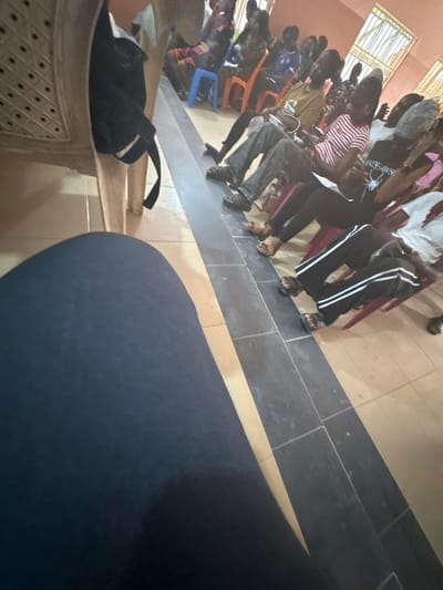The scene depicts an indoor gathering in Iriebe, Nigeria, observed from a first-person, seated perspective. In the lower left foreground, the dark blue/black fabric of the camera operator's leg is visible. To the immediate left, a beige plastic chair holds a black backpack. The floor features light-toned tiles in the foreground, transitioning to dark grey/black tiles further back, separated by a distinct black tiled strip running diagonally from mid-left to lower right. Approximately 15-20 individuals, primarily young adults, are seated in multiple rows on assorted plastic chairs (blue, orange, red, beige) in the midground and background. They are dressed in casual attire, including various t-shirts, pants, and sandals. Several individuals hold white papers or small objects, possibly mobile phones, and are generally facing forward, suggesting a collective focus on an unseen speaker or presentation area. The background wall is light-colored, with grid-patterned windows admitting natural light from the upper right.