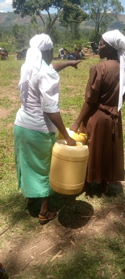 Two women, both wearing white headscarves, stand in a grassy field, possibly in Malava, Kenya, at the coordinates provided. The woman on the left is wearing a white shirt and a teal skirt, and she is holding a large yellow plastic container. The woman on the right is wearing a brown dress and is holding a smaller yellow plastic container. Both women are looking off-camera to the right. The ground is covered in green grass and scattered pieces of dried branches.  The background features a group of people sitting in the distance, a few motorcycles, and trees. The weather is sunny and clear.