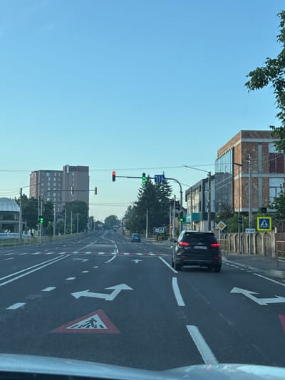 The image shows a street scene in Vatra, Moldova, on a clear day with a blue sky. The perspective is from inside a vehicle looking forward. The road is paved and has multiple lanes with white lane markings and a red and white crosswalk marking on the ground. There are traffic lights visible, some showing green and one showing red. A black SUV is driving in the right lane, and a blue car is further ahead in the left lane. Buildings of various sizes and architectural styles line the sides of the road, including a tall, multi-story building with many windows in the background. Trees and streetlights are also present. A "STOP" sign and a pedestrian crossing sign are visible on the right side of the road. The overall environment appears to be a typical urban or suburban street with moderate traffic.