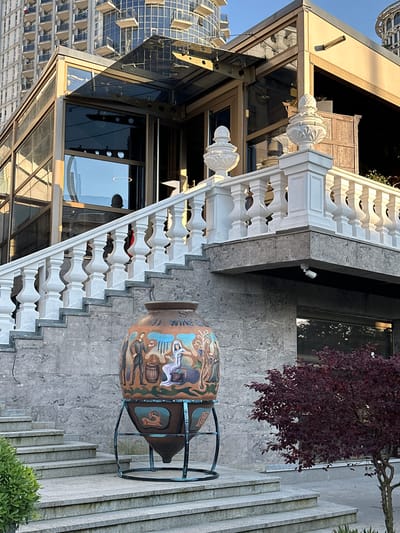 This image captures an outdoor urban scene in Batumi, Georgia, featuring a building, a staircase, and a large decorative vessel.

In the foreground, a large, ovoid-shaped clay vessel, consistent with a traditional Kvevri, rests on a dark metal stand. The vessel is decorated with colorful painted scenes depicting figures engaged in activities related to wine production, and the partial inscription "WINE GA" is visible near its top. To the left of the vessel, a multi-tiered stone staircase ascends. A small tree with dark red foliage is planted to the right of the vessel, and a small green bush is partially visible on the far left.

The stone staircase leads up to a white balustrade with decorative finials, indicating a raised terrace or balcony. A white security camera is mounted underneath the edge of this elevated structure. The main building behind the balustrade features a modern architectural design with light-colored walls and extensive glass paneling. Reflections in the glass show segments of the sky, adjacent buildings, and at least two discernible human silhouettes. A clear glass canopy extends from the building's upper section.

In the background, several modern high-rise buildings are visible. One prominent structure is a light-colored tower with numerous uniform balconies. Another distinct building exhibits a curved glass exterior. The scene is illuminated by natural daylight, producing both highlights and shadows.