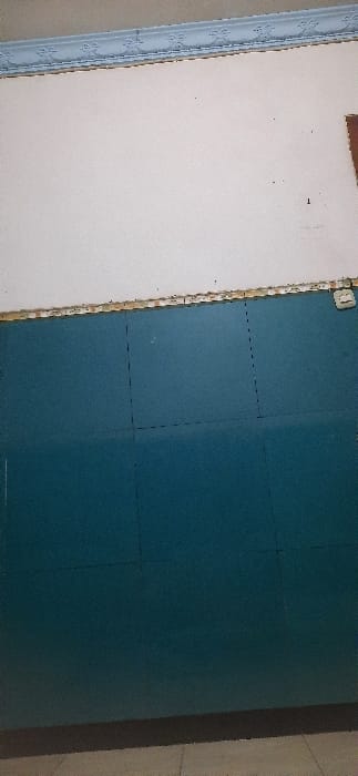 The image shows the interior of a room, possibly a school classroom or office. The walls are painted, the top half is white and the bottom half is teal. There are no people present, and the room is empty except for the walls. There are two thin, black vertical lines dividing the teal wall into three sections. The room has a white ceiling with a decorative, slightly ornate molding. The floor is a light brown tile. There is a small white square object on the right side of the wall near the edge of the image. The coordinates -7.488, 112.715 place the location in the city of Kecamatan Candi, Indonesia.