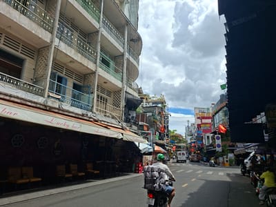 The image depicts a street scene in Thủ Đức, Vietnam, on a cloudy day. A multi-story building with balconies lines the left side of the street. A sign on a storefront awning reads "Lucky bar". In the foreground, a person rides a motorcycle loaded with packages. The street is paved, with other vehicles, including cars and motorcycles, visible further down the road. The overall atmosphere suggests a bustling urban environment.