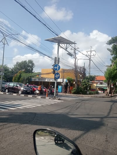 The image captures a street scene in Surabaya, Indonesia, focusing on a busy intersection with a prominent "WARKOP AMPIRO" sign on a yellow building. The foreground shows a motorcycle's side mirror reflecting the road and other vehicles. In the middle ground, a pedestrian crossing is visible, with several cars and motorcycles stopped at a traffic light. Traffic signs indicating left and forward turns are mounted on a pole alongside the traffic signal. The background features a tangle of electrical wires overhead, typical of urban Indonesian streetscapes, and lush green trees bordering the street. The sky is a mix of blue with scattered white clouds. People on motorcycles are seen wearing helmets, indicating adherence to traffic safety regulations. The overall scene depicts a typical day at a bustling urban junction in Surabaya.