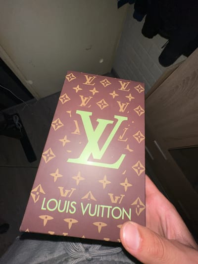 A close-up, eye-level shot shows a person's right hand holding a brown Louis Vuitton branded box with a repeating pattern of the Louis Vuitton monogram and flowers. The box has large, light green "LV" lettering and "LOUIS VUITTON" text. The background is dark and out of focus, showing a wooden cabinet and a wall with a subtle brick pattern. No specific location within Kazanlak, Bulgaria is discernible from the image.