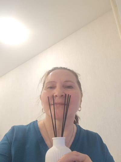 A woman is holding a white, ridged vase filled with black diffuser sticks in front of her face. She is wearing a teal V-neck shirt and a silver necklace. The background consists of a plain, off-white wall and a ceiling light fixture. The photograph appears to have been taken indoors. There is no verifiable information to determine the location in the city of Sarapul, Russia.