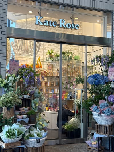 The image shows the storefront of "Kate Rose," a flower shop located in Chiyoda, Japan. The facade features large glass doors and windows, revealing a well-lit interior decorated with a brick-patterned wall. The shop's name, "Kate Rose," is prominently displayed in silver, three-dimensional lettering above the entrance.

Inside, the shop is filled with an abundant display of flowers and plants. Various arrangements are showcased, including large bouquets of blue hydrangeas and purple wisteria-like flowers in the foreground. Baskets filled with white and green flowers are placed on wooden crates, and smaller potted plants and decorative vases are visible on shelves. In the background, framed dried flower arrangements are displayed.

A person, wearing a dark hooded jacket and jeans, is working inside the shop, appearing to arrange flowers. They are positioned behind a counter, with their back to the viewer.

The exterior of the shop is lined with light-colored brickwork, and the entrance has a modern, metallic frame. The overall impression is of a professional and aesthetically pleasing floral business.