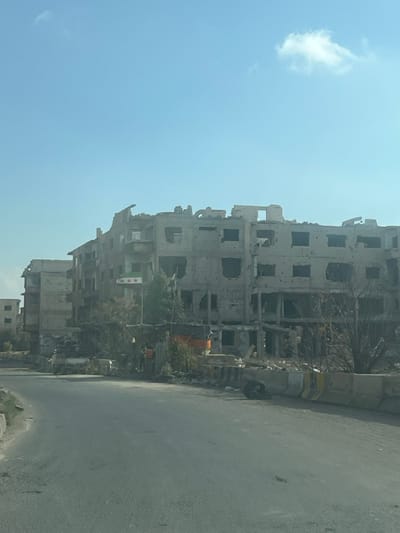 A heavily damaged building stands prominently, marked with numerous bullet holes and structural decay. A Syrian flag hangs from it. The road in front of the building is clear and smooth. A tire rests near the road. A single cloud floats in the light blue sky. The image suggests urban destruction, likely due to conflict or war.