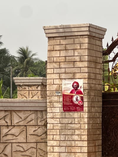 The image displays a light beige, rectangular brick pillar located in Nnobi, Nigeria, serving as a support for an ornate dark metal gate on the right. A reddish-brown "Glorious Exit" funeral announcement poster is affixed to the pillar. The poster features a smiling older woman in a red headwrap and traditional attire, identified as "EZINNE LADY EUNICE UGOYE MBAKWE," above a circular inset image of an older man in a traditional hat, positioned next to a red numeral "91." Below the poster, a white rectangular sign states "AMADUNU HOUSE" and "121." The pillar's cap shows visible cracks. To the left of the main pillar, a lower stone wall with carved patterns extends, beyond which dense green tropical vegetation, including multiple palm trees, is visible against a bright, overcast sky. No individuals or active interactions are visible in the scene.