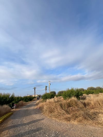A landscape scene, possibly in the Saâda region of Morocco, features a dirt road bordered by dry, grassy vegetation and interspersed with green bushes and trees. Overhead, the sky is partly cloudy with streaks of blue. Utility poles are visible along the roadside. A parked motorcycle with a black figure in close proximity can be seen on the lower right.