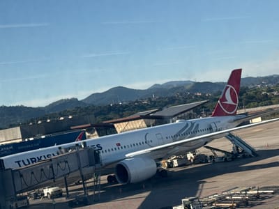 A Turkish Airlines Airbus A330-300 aircraft, identifiable by registration TC-JJR on its red tail fin, is parked at a gate on the tarmac of São Paulo/Guarulhos International Airport (GRU) in Guarulhos, Brazil. A jet bridge is connected to the forward left passenger door, and a mobile passenger stair is positioned at the rear left door. Ground support equipment, including a pushback tug, baggage carts, and various service vehicles, is arranged around the aircraft. Several ground crew personnel are visible on the tarmac and near the jet bridge. A blue LATAM Cargo aircraft, bearing the text "LATAM CARGO PERISHABLE HUB," is partially visible behind the Turkish Airlines jet. Airport buildings with angular roofs are visible in the mid-ground. The distant background features rolling, verdant hills and mountains, with scattered residential areas visible on their slopes, beneath a clear, light blue sky containing several faint contrails. The scene is illuminated by bright sunlight, suggesting daytime operations.