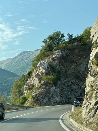 The image depicts a scenic mountain road in Košljun, Montenegro. A black Volkswagen Golf is parked on the right side of the road, partially obscured by a rocky cliff face. The road curves to the right, with a white line marking the lane separation and a curb on the right edge. Vegetation, including trees and bushes, grows along the rocky slopes of the mountain. In the background, rolling green hills and mountains stretch towards a clear blue sky with scattered white clouds. The overall scene is one of natural beauty and tranquility, with the road appearing to be a well-traveled route through the mountainous terrain.