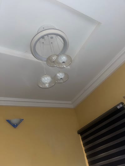 The image shows an interior view of a room with a yellow wall and a white ceiling. A modern chandelier with a circular frame and four hanging glass lights is suspended from the ceiling. On the yellow wall, there is a triangular blue light fixture with a white diffuser. A portion of a black and grey striped roller blind is visible on the right side of the image. The scene is indoors and appears to be well-lit, likely in a residential or commercial setting within the city of New Stadium, Nigeria. There are no people visible in the image.