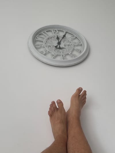 The image shows a white, ornate wall clock with Roman numerals against a plain white background. The clock face has intricate scrolling patterns. The time displayed on the clock is approximately 10:07. To the right of the clock, two bare feet and lower legs of a person are visible, with the legs crossed. The person's legs are hairy. The overall scene is minimalist and appears to be indoors, possibly in a home or studio setting. There is no visible text in the image, and no specific cues about the time of day, weather, or emotions are apparent. The setting is identified as Molenbeek-Saint-Jean - Sint-Jans-Molenbeek, Belgium.