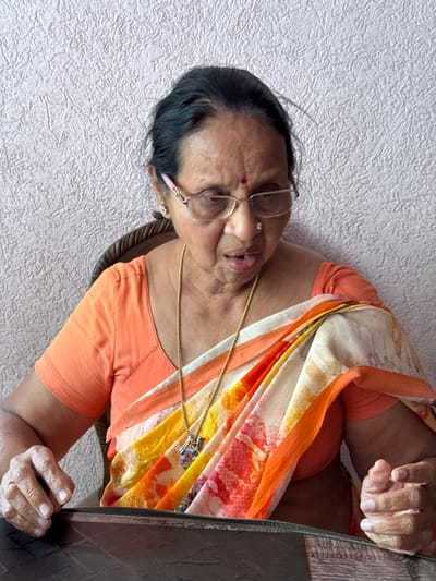 An elderly woman wearing glasses and traditional Indian clothing sits in a chair. Her sari is a vibrant mix of orange, white, and floral patterns. She has a gold necklace and a small nose piercing. The background includes a textured, light-colored wall and the edge of a dark, possibly wooden, table or surface. Her hands are near the table's edge.