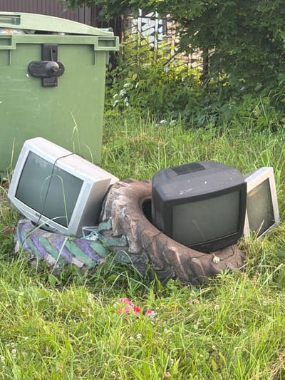 The image depicts an outdoor scene in Красный Маяк, Russia, featuring discarded electronics and a trash bin amidst lush green grass and foliage. Two older model televisions are prominently visible. One is a black, bulky CRT television partially nestled within a large tractor tire that has a distinctive tread pattern and is decorated with green and purple stripes. Another, lighter-colored television, also appears to be a CRT model, is placed behind and slightly to the left of the black TV.
A large, light green trash bin is positioned on the left side of the frame. Its lid is partially open, and a black latch mechanism is visible on its side. In the background, dense green leaves and branches of trees suggest a natural, perhaps slightly overgrown, environment. A fence with a diamond pattern is partially visible through the foliage. The overall impression is one of abandonment and waste disposal, with the electronic items and the tire likely awaiting collection or being improperly discarded.
The lighting and the vibrancy of the grass suggest it is daytime, likely with fair weather. There are no people visible in the image. The scene appears to be a somewhat neglected outdoor area, possibly a yard or a designated waste collection spot. No text is discernible in the image.