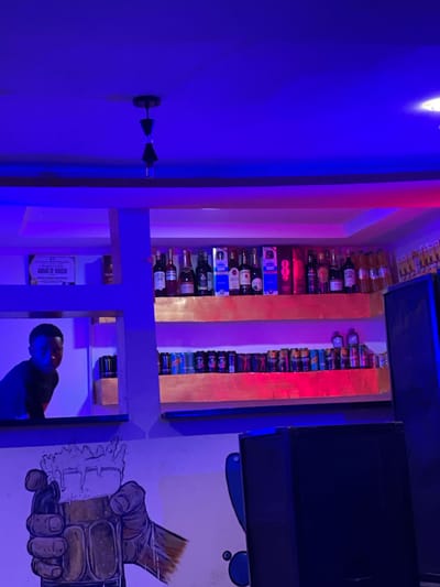 A man is present in a dimly lit room with blue and pink lighting. The room appears to be a bar or a lounge, with shelves displaying various bottles of alcohol. The shelves are made of wood and are illuminated by colored lights, creating a vibrant atmosphere. In the background, there is a mural on the wall depicting a hand holding a frothy drink. The man, who is partially visible in the frame, seems to be looking away from the camera. There is text visible on a certificate or award hanging on the wall, which reads "AWARD OF HONOUR" and "SQUAD". Other visible text includes brand names such as "CAMPARI", "JAMESON", and "PM". The overall impression is that of a lively, modern establishment. The time of day is likely evening or night due to the artificial lighting.