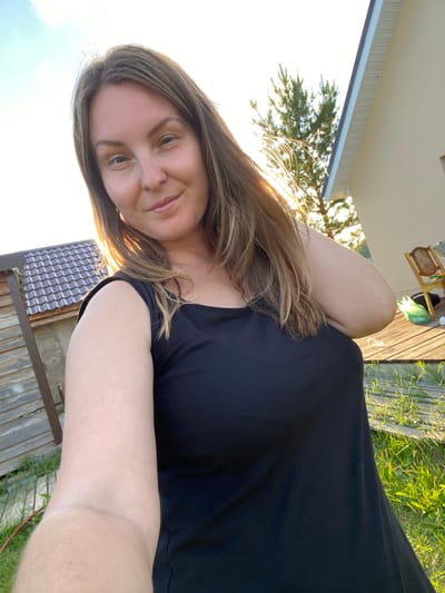 A woman with long, light brown hair and fair skin takes a selfie outdoors. She is wearing a black, sleeveless top and is standing on a wooden deck with green grass and a wooden shed in the background. Sunlight is casting a warm glow on her face and hair. To the right of the frame, part of a light-colored house and a tall green tree are visible. The scene appears to be in a rural or suburban setting, possibly in or near the village of Garevaya, Russia, indicated by the presence of a shed, wooden structures, and natural greenery.