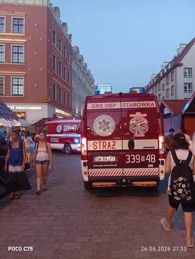 This media file captures a street scene in Wrocław, Poland, likely in the evening given the ambient lighting and the timestamp. The primary subjects are two ambulances, identified as belonging to "GRS OSP STARÓWKA," parked on a cobblestone street. One ambulance is a bright red, bearing the markings "STRAŻ" (meaning "Fire Department") and "AMBULANS." It displays a license plate "DW 8AJ71" and the number "3390-48." The other ambulance, partially visible, also shows similar red livery and has "3390-47" on its side.
In the foreground, two women are walking on the cobblestone pavement. The woman on the left wears a blue and white tie-dye dress and sandals. The woman on the right wears a light-colored, lacy top and denim shorts, carrying a black backpack adorned with various patches, including some associated with the Los Angeles Lakers.
The background features buildings with traditional European architecture, characteristic of historic city centers. One building, painted in a reddish-orange hue, has large windows and ornate detailing, with illuminated signage reading "VN PIEROGARNIA" and "STARY MŁYN PIEROGARNIA," indicating food establishments. The sky is a pale blue, suggesting either early evening or a bright twilight. The overall atmosphere appears calm, with no overt signs of urgency, despite the presence of emergency vehicles. The timestamp "26.06.2026 21:33" and the text "POCO C75" are also visible in the lower portion of the image.