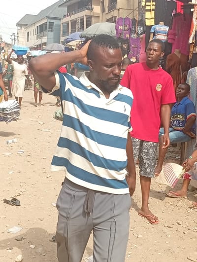 The image depicts an outdoor scene on an unpaved, sandy street in Fegge, Nigeria, characteristic of a market environment. The ground is light brown and uneven, with scattered debris including paper and plastic items. Multi-story concrete buildings line the background, interspersed with market stalls displaying various hanging garments in colors such as purple, red, yellow, and blue. Utility poles and wires are visible in the distance.
In the foreground, slightly off-center, a dark-skinned adult male stands, facing generally forward but looking downward and to his right. He wears a cream and blue horizontal striped polo shirt with a dark emblem on the left chest, and grey track pants with a visible drawstring. His right hand is placed on the back of his head/neck. He has short, dark hair and a beard.
To the right of the main subject, a dark-skinned young male stands, wearing a red short-sleeved t-shirt with a circular emblem on the left chest and black and white patterned shorts. He looks towards the left side of the frame. Further back on the left, a dark-skinned woman in a light-colored dress walks away from the camera, balancing a blue basin on her head. On the far right, partially visible, a person is seated, wearing a blue shirt and holding a woven fan with red and white stripes. Their bare feet, adorned with red flip-flops, are visible on the ground. The scene is brightly lit by natural sunlight.