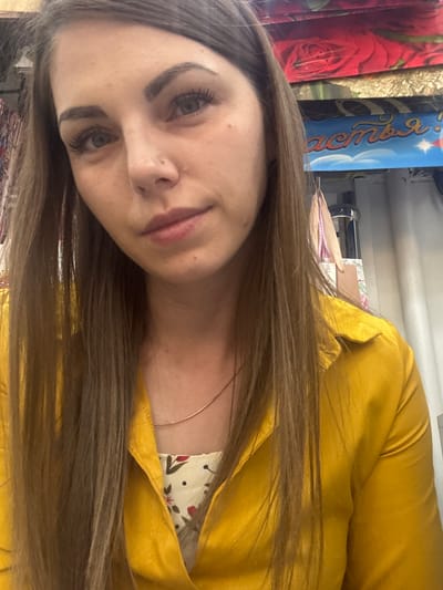 A selfie-style photo shows a woman with light skin, brown hair, and green eyes. She wears a yellow garment over a floral top and a thin gold necklace. The background includes fabric rolls with floral patterns and a blue cylindrical object with Russian lettering. The environment suggests a fabric store setting, possibly located in Klin, Russia, based on the Cyrillic characters on the blue object.
