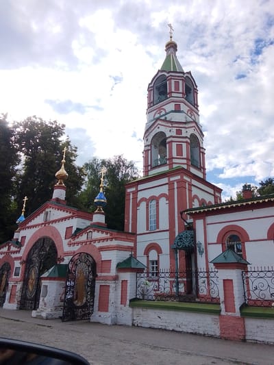 The image shows a frontal view of a Russian Orthodox church and its adjacent bell tower, located in the city of Staryi Pokrov, Russia. The architecture is characterized by white brick walls with red decorative elements and multiple golden domes topped with crosses. The bell tower is tall and octagonal, with arched openings and a green conical roof. In the foreground, there is an ornate wrought-iron gate with decorative elements and golden icons visible behind it. A person is seen standing behind the open gate. The sky is partly cloudy. The environment suggests a sunny day.