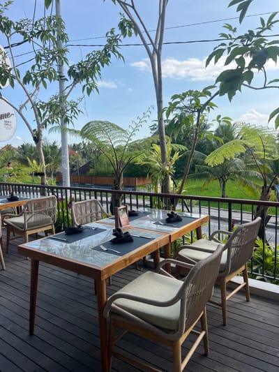 The image shows an outdoor dining area in Tegallalang, Indonesia, with a view of lush greenery and rice paddies. In the foreground, a wooden table with a glass top is set for dining, complete with placemats, cutlery, and folded napkins. Several woven chairs with light-colored cushions surround the table, which is placed on a dark wooden deck. Beyond the railing of the deck, a vibrant green rice field stretches out, bordered by trees and a building. The sky above is blue with scattered white clouds. The overall scene suggests a peaceful and scenic dining experience.