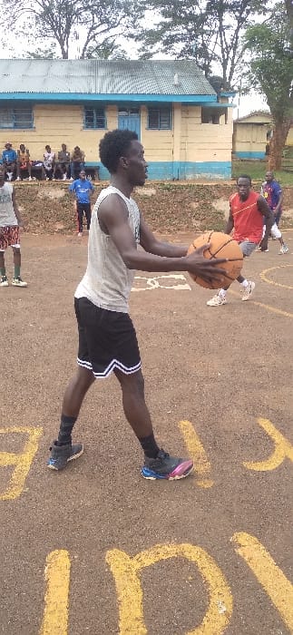 A group plays basketball on an outdoor court. A man in a grey tank top and black shorts with white trim is about to shoot. He holds the basketball above his head. A man in a red shirt is standing behind him to his right. Spectators sit or stand in the background. The court is painted with yellow markings. A building with a light-yellow exterior and a blue trim and roof is also visible in the background.
