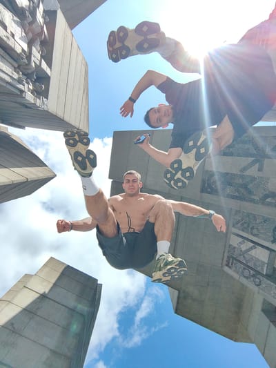 Two young men are engaged in parkour on a sunny day, beneath a clear blue sky with some scattered clouds, at the Buzludzha monument, a former socialist party house located in Shumen, Bulgaria. The camera angle is from below, looking up at the men. One man, in the foreground, is shirtless and wearing dark shorts and white socks with athletic shoes. He is in mid-air, with his legs bent and one foot extended upwards. His left arm is extended downwards, and his right arm is bent, with his fist clenched. He has a tattoo on his left arm. The second man is in the background, wearing a dark t-shirt and dark shorts. He is also in motion, with one leg bent and the other extended. He is holding a mobile phone in his right hand. The monument's concrete structure dominates the background, with its angular and imposing architecture visible against the sky. The overall impression is one of athletic activity and urban exploration in a distinctive architectural setting.