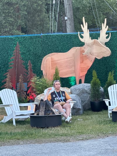 A person sits in a white Adirondack chair next to a fire pit at an outdoor event. The person is wearing a black and orange t-shirt and white sneakers, with sunglasses perched on their head. They are holding a small orange object in their right hand. Behind them, a large wooden cutout of a moose stands against a green backdrop, with metal cutouts of trees to the left. Two other white Adirondack chairs are visible on either side of the central scene. The ground is covered in grass and gravel. The overall scene suggests a festival or outdoor gathering. The location is not specified in the image, nor does the image provide any clues to determine it is in Vancouver, Canada.