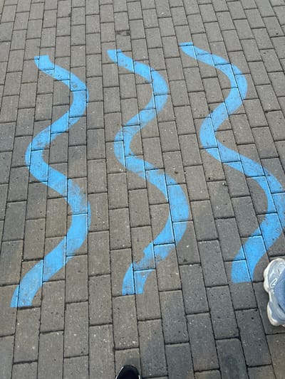 The image shows a pedestrian walkway paved with gray rectangular bricks. Three wavy blue lines are spray-painted on the pavement, running diagonally from the upper left to the lower right of the frame. The lines appear to be markings for a path or a designated area. A person's legs, wearing gray athletic shoes and blue jeans, are visible in the lower right corner of the image, standing on the walkway. The lighting suggests it is daytime. The scene is a public outdoor space, consistent with an urban environment.