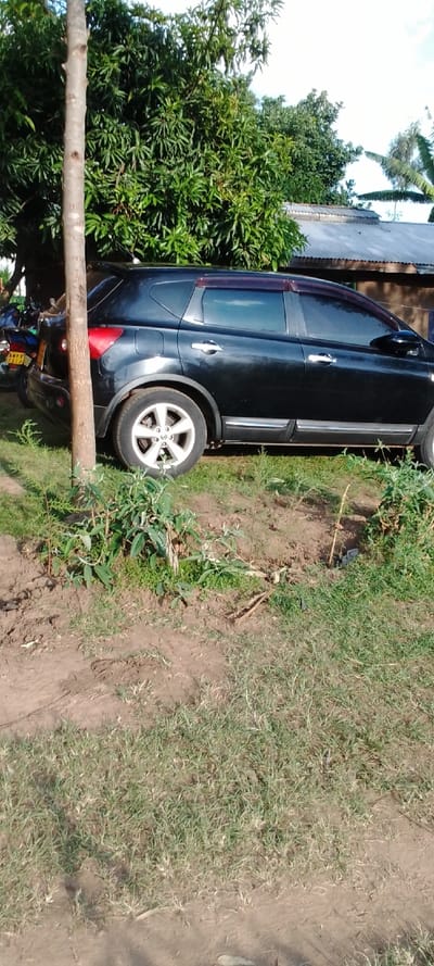 A black SUV is parked on a dirt path next to a tree trunk in Kipkarren, Kenya (latitude: 0.599, longitude: 34.957). The vehicle is parked on the edge of a small, grassy area. There is a small, brown building behind the vehicle.  The SUV is parked facing the right, and its front wheels are turned slightly to the left. The tree trunk is in the lower left portion of the image.  The ground is dry and dusty.  There are other trees in the background.  There are no people in the image.  The image is taken at a low angle.