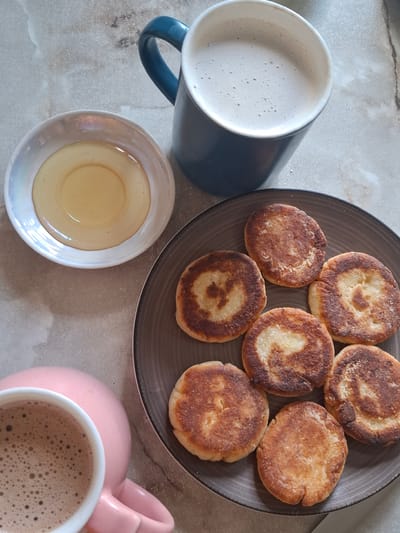 The image depicts a breakfast scene on a neutral-toned, textured surface. In the upper right, a dark blue mug filled with a frothy, light brown beverage, likely coffee or hot chocolate, is visible. To the left of the mug, a small, iridescent white saucer holds a light golden liquid, appearing to be honey. In the foreground, a pink mug with a similar frothy, light brown beverage is partially visible on the left. Dominating the center and right of the frame is a dark gray plate containing a circular arrangement of six golden-brown, fried pancakes or fritters, each with slightly browned edges and lighter centers. The overall composition suggests a home-prepared meal, with no discernible elements indicating a specific location in Donetsk, Ukraine, or any interaction between individuals.