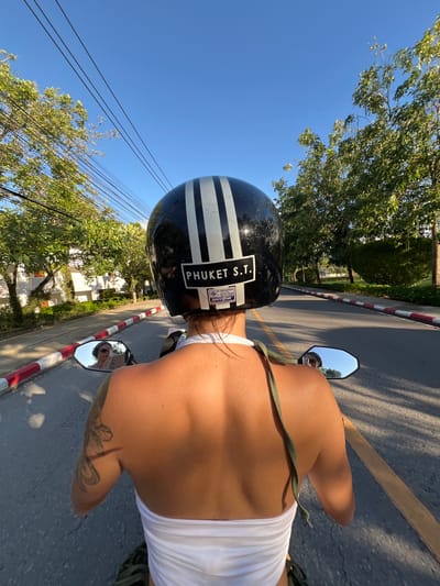 A person wearing a black and white striped helmet with "PHUKET S.T." written on it is riding a motorcycle. The person is wearing a white halter top and has a tattoo on their left arm. The motorcycle has a rearview mirror that reflects the rider's face. The rider is on a road with red and white striped borders. Trees line the side of the road, and there are buildings in the background. The sky is clear and blue. The location is Choeng Thale, Thailand.