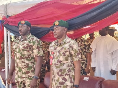 The image shows a group of people, primarily men, standing under a canopy. The canopy is made of red, black, and white fabric. The men are wearing military uniforms, with the exception of one man who is dressed in a white robe with a cape. The location is the city of Maiduguri, Nigeria, based on the provided coordinates. The men appear to be attending a ceremony or event, and the occasion is unclear. The men's facial expressions are mostly stoic, with a few smiling or laughing. The environment is outdoors and appears to be sunny.